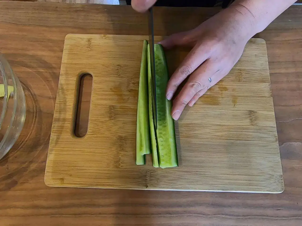 Ręka kroi ogórka na desce, przygotowując go do sushi. Pokrojone paski ogórka są gotowe do użycia.