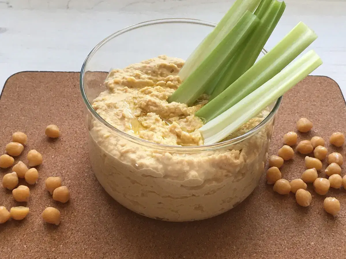 Pyszny hummus z ciecierzycy w szklanej misce, z łodygami selera do maczania i rozsypanymi ziarnami ciecierzycy na blacie.