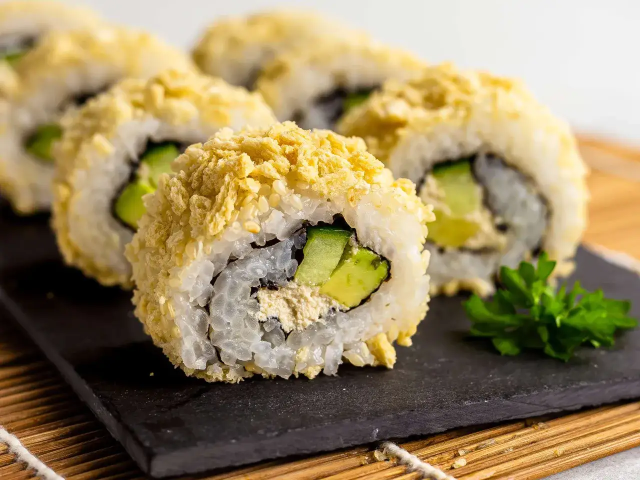 Chrupiące rolki sushi w panko z awokado i ogórkiem, podane na czarnej desce, ozdobione natką pietruszki.