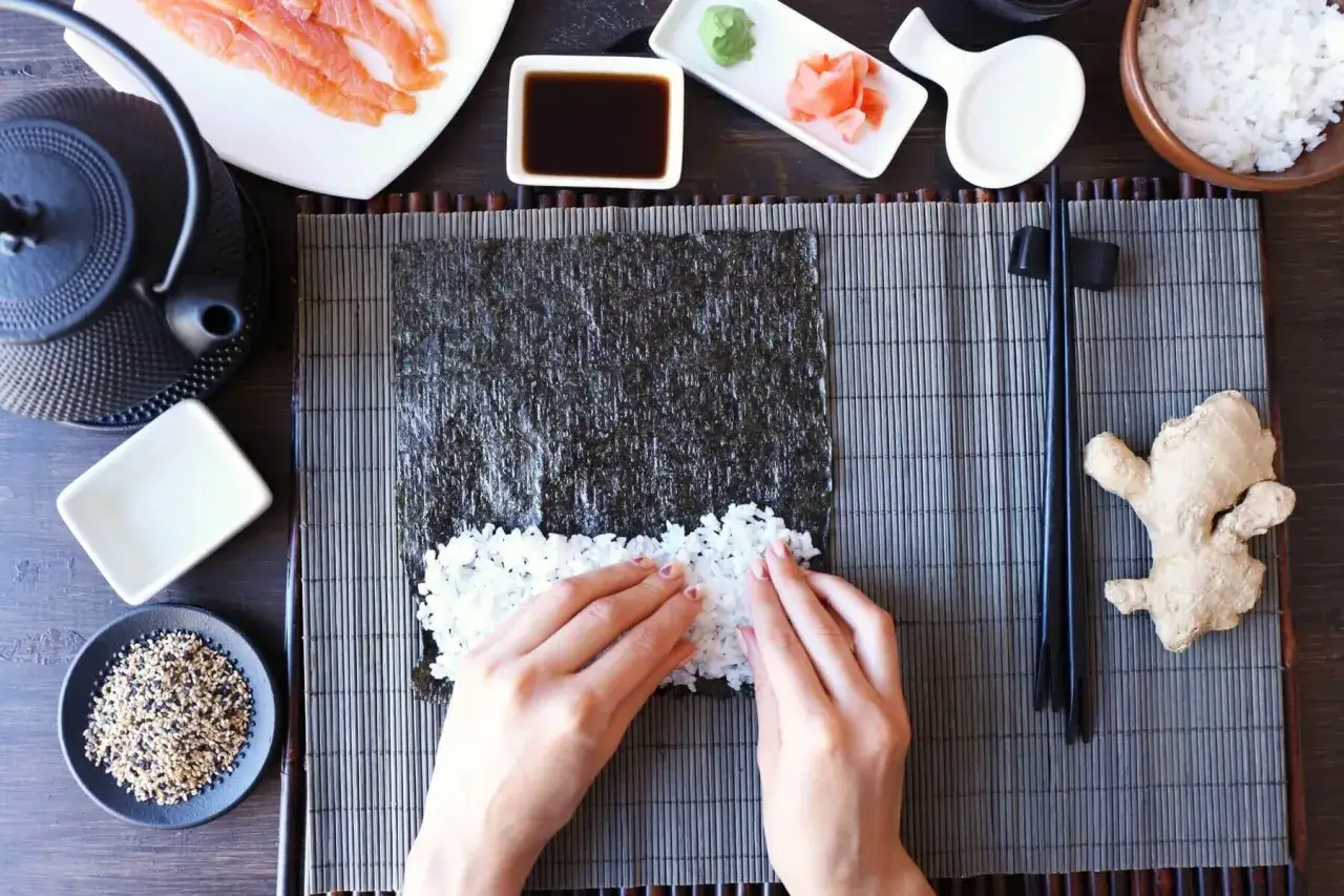 Ręce rozprowadzają ryż na nori, przygotowując do zwinięcia sushi. Obok łosoś, sos, imbir i ryż. Zaprawa do sushi przepis w zasięgu ręki.