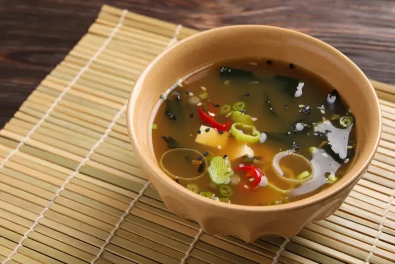 Aksamitna zupa miso z tofu, wodorostami, szczypiorkiem i papryczką chili, podana w ceramicznej miseczce na bambusowej macie.